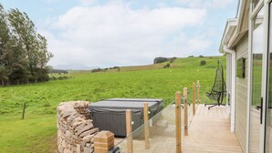 Une terrasse en bois mène à une maison dont un des murs est en pierre et qui possède un jacuzzi au centre.