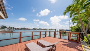 Eine Holzterrasse mit zwei Liegestühlen und einem Tisch, mit Blick auf ruhiges Gewässer, eine entfernte Küstenlinie und Palmen.