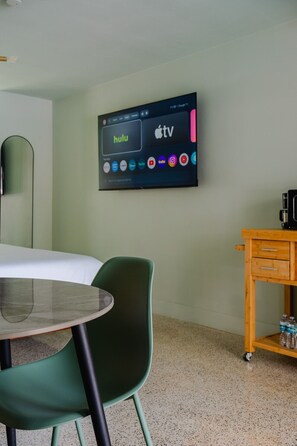 Habitación de hotel con una cama, una silla verde, una mesita pequeña, un televisor montado en la pared que muestra servicios de streaming y una mesita de noche de madera con botellas.