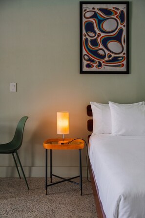 Habitación de hotel con cama, mesita de noche con lámpara, una silla verde y un cuadro abstracto colorido en la pared.