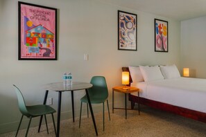 A hotel room with a bed, a table with chairs, a water bottle, and two framed artworks on the wall.