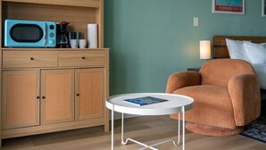 Una habitación con un armario de madera, un microondas azul, una mesa redonda blanca, un sillón marrón y una cama con almohadas blancas.