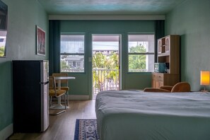 Une chambre d’hôtel comprenant un lit, un bureau, une chaise, un réfrigérateur, une armoire, une lampe et un balcon avec vue sur les bâtiments et la verdure.