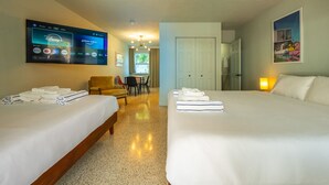 Ein Hotelzimmer mit zwei Betten, einem Fernseher, der Streamingdienste anzeigt, einer Sitzecke mit Sessel und Tisch sowie einer Tür zu einem weiteren Raum.
