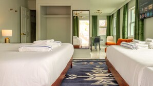A hotel room with two beds, a desk, and a chair. There is a window with curtains, a lamp, and a wall-mounted screen displaying app icons.
