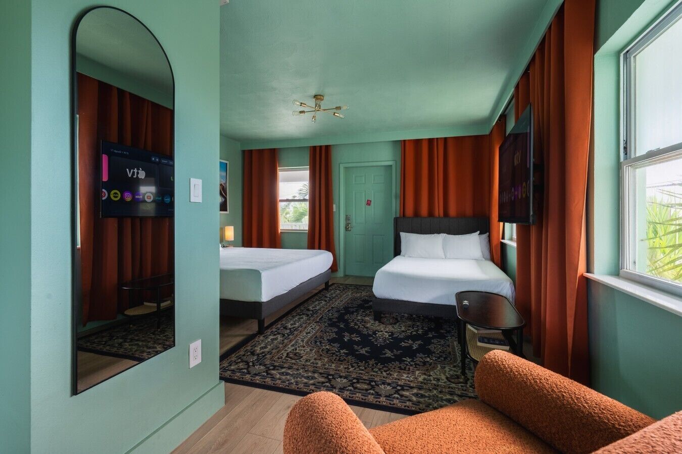 A hotel room with two beds, a large mirror, a television, and a window with curtains.