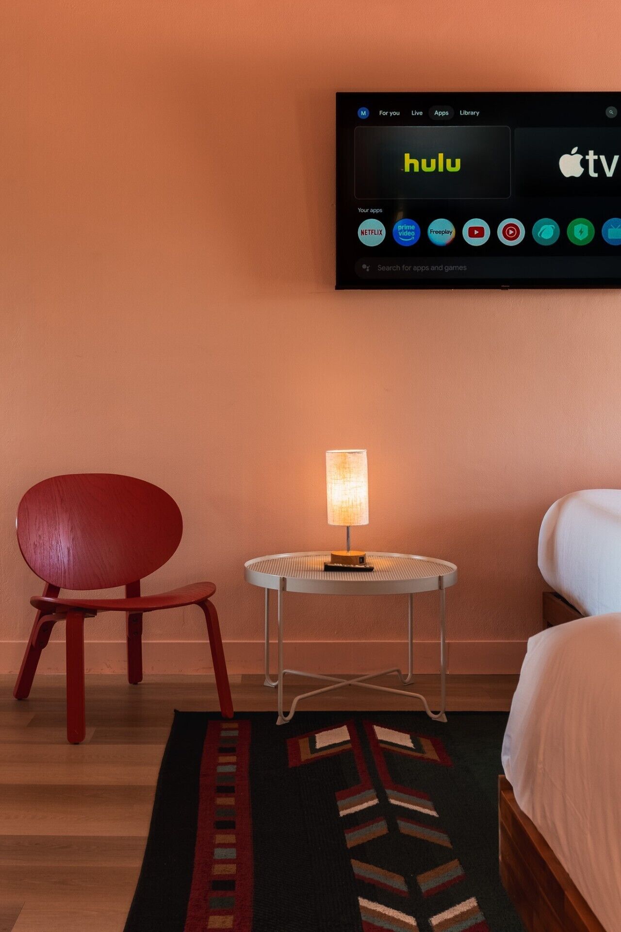 A hotel room with a television mounted on the wall displaying Hulu, a red chair, a small table with a lamp, and a bed with white bedding.