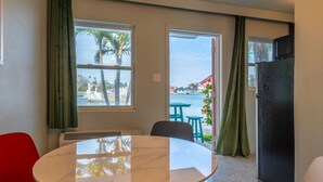 A room with a round table, two chairs, a black refrigerator, and a view of a beach with palm trees and a pool.