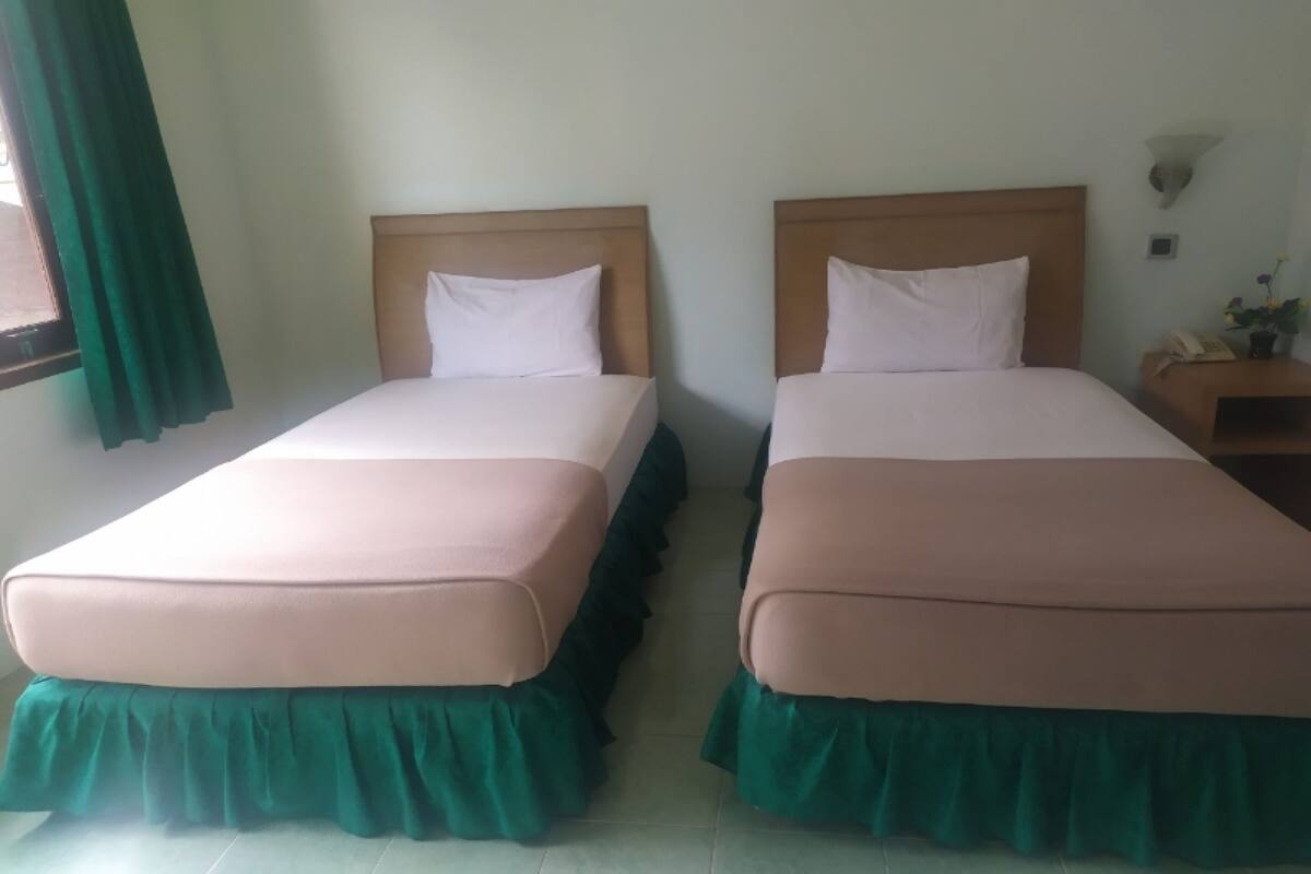 Dos camas individuales con cabeceros de madera y cubrelechos verdes, una mesita de noche con lámpara, y una ventana con cortinas verdes.