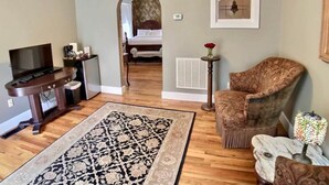 Un salon avec un plancher en bois, un tapis à motifs, un téléviseur, une machine à café, une petite table avec un vase et un fauteuil.