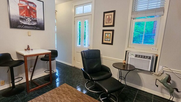 A dining area with a table and two chairs, a black leather chair, a small round table with a decorative item, and a window with blinds. - The Moderne Suite Downtown Balcony, Parking (Eureka Springs)