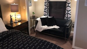 Ein kompaktes Hotelzimmer mit Bett, einer schwarzen Ledersofa, einem kleinen Tisch mit Lampe und gemusterten Vorhängen.