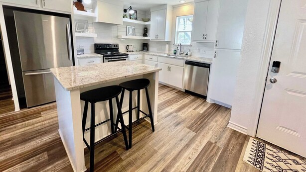 A modern kitchen with white cabinetry, a stainless steel refrigerator, a built-in oven, and a dishwasher. There are two black bar stools at the island. - Serenity Stay - 3BR 2BA - South Lubbock (Lubbock)