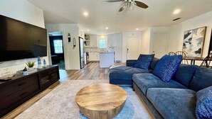 A modern living room with a large flat-screen TV, a wooden coffee table, a gray sofa, and a ceiling fan. - Serenity Stay - 3BR 2BA - South Lubbock (Lubbock)
