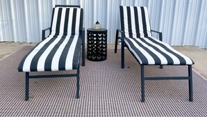 Two black and white striped lounge chairs with a small round table between them, set on a woven outdoor floor, against a corrugated metal wall. - Serenity Stay - 3BR 2BA - South Lubbock (Lubbock)