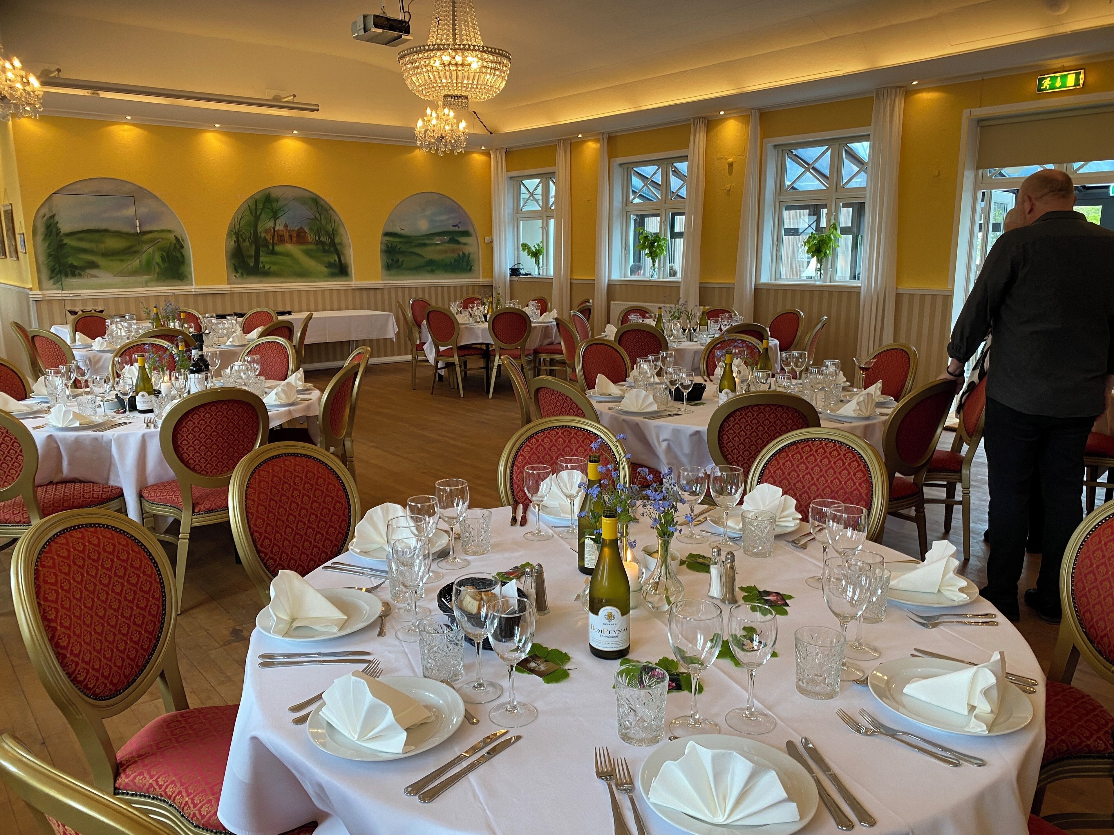 A formal dining room with round tables, red upholstered chairs, and chandeliers.