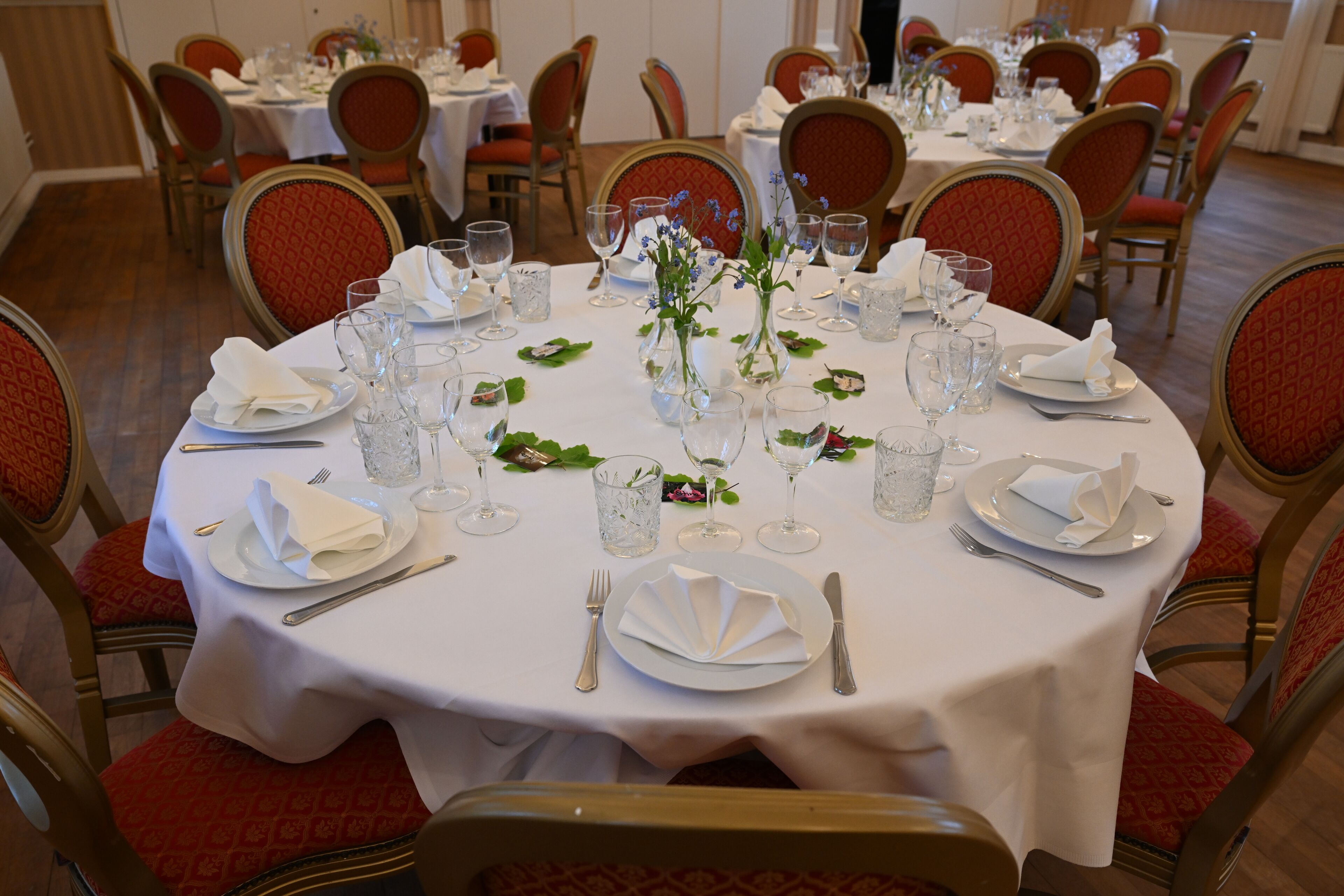 Un comedor con mesas redondas dispuestas para cuatro personas, cada una con manteles blancos, sillas tapizadas en rojo y una vajilla ordenada que incluye platos, cubiertos y copas.