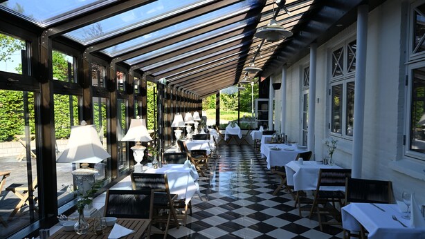 A covered outdoor dining area with checkered flooring, tables set with white tablecloths and chairs, and large windows on one side. - Rold Gl. Kro (Arden)