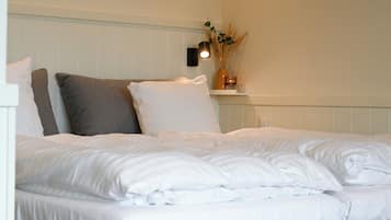 A neatly made bed with white linens and pillows, a bedside lamp, and a small shelf with a vase of dried plants.
