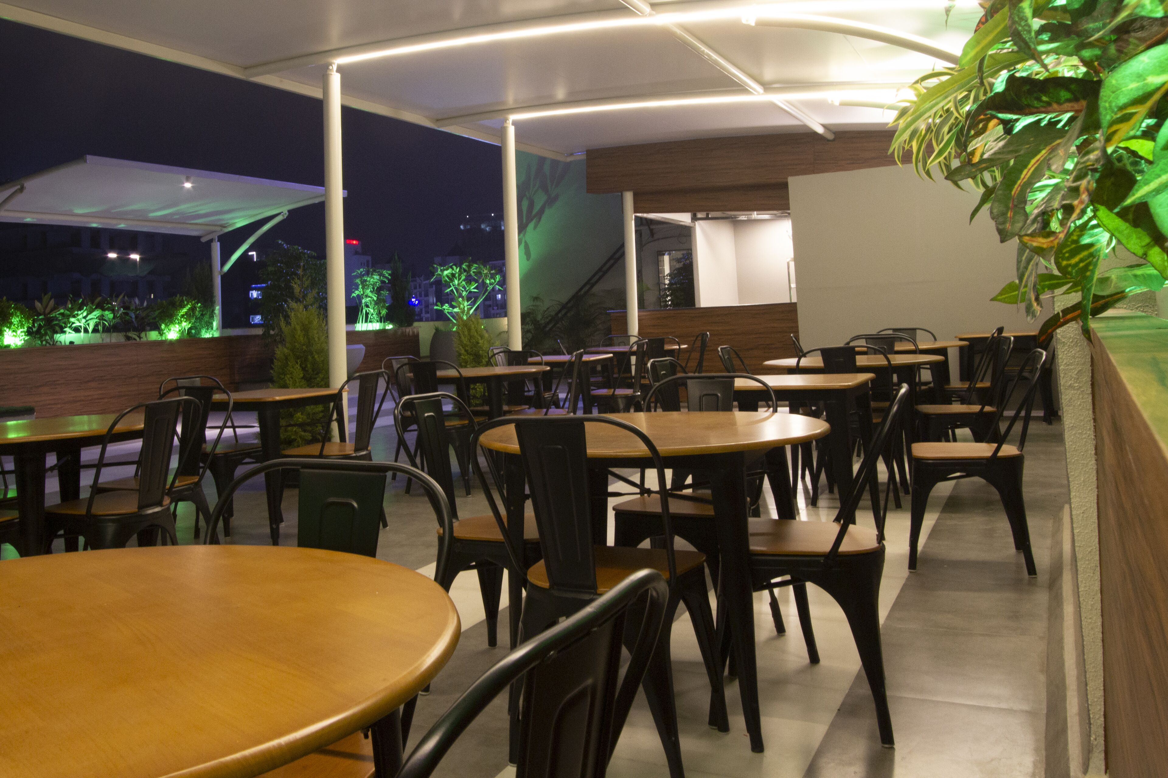 An outdoor dining area with multiple tables and chairs, surrounded by plants and a covered walkway.