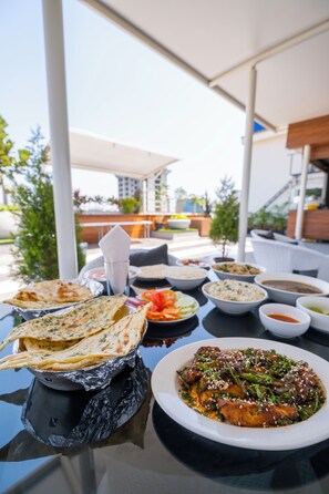 A table with various dishes, including flatbreads, rice, and a fish dish, set outdoors with a view of buildings and greenery in the background. - Monarch Guestline Magarpatta (Pune)