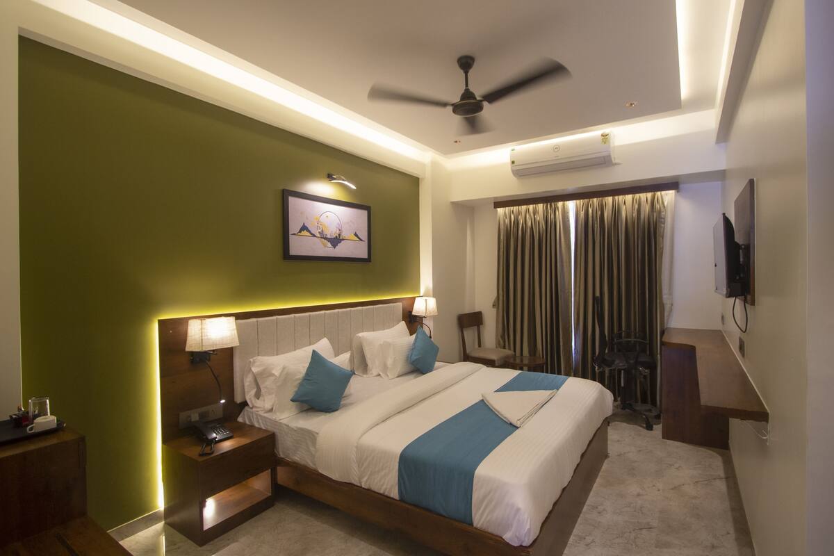 A hotel room with a bed, bedside tables, a desk, a chair, a ceiling fan, and a TV on the wall.