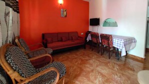 Uma sala de estar com parede vermelha, um sofá, duas poltronas, uma mesa de jantar com cadeiras, um ventilador de teto e uma televisão.