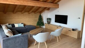 Ein modernes Wohnzimmer mit Holzboden, einem großen Flachbildfernseher, einer Eckcouch, zwei Sessel und einem kleinen Weihnachtsbaum.