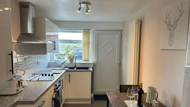 A modern kitchen with a central island, a dining table with chairs, and a view of the outdoors through a window. - Immaculate 1-bed House in Newtown, Disley (Stockport)