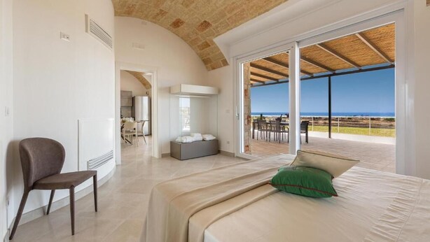 Un dormitorio con una cama grande, una silla y vista a la playa a través de una puerta de cristal.