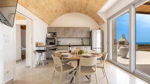 Una cocina moderna con techo abovedado, un comedor con mesa dispuesta para cuatro personas y vista a la playa a través de amplios puertas correderas de cristal.