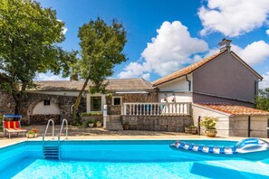A swimming pool with a clear blue water surface, surrounded by a stone wall, a wooden deck, and a building with a tiled roof. - Meja Stone House With Pool (Bakar)