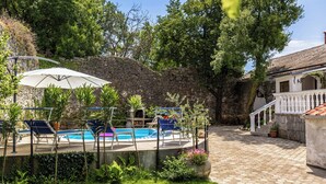 Area piscina con pareti in pietra, poltrone relax e ombrellone bianco.