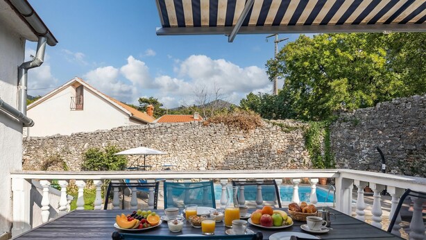 Area pranzo all'aperto con tavolo apparecchiato per la colazione, una piscina, pareti in pietra e vista su una collina.