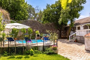 A pool area with lounge chairs and umbrellas, surrounded by greenery and stone walls. - Meja Stone House With Pool (Bakar)