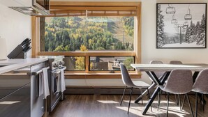 Una cocina moderna con un ventanal amplio que da a un área boscosa, un comedor con sillas, y una isla de cocina con estufa y horno.