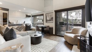 Una sala de estar moderna con sofá, sillón, mesa de centro y comedor con sillas y una mesa. Se ve una cocina al fondo, un ventanal con vistas a árboles y una lámpara en el techo.