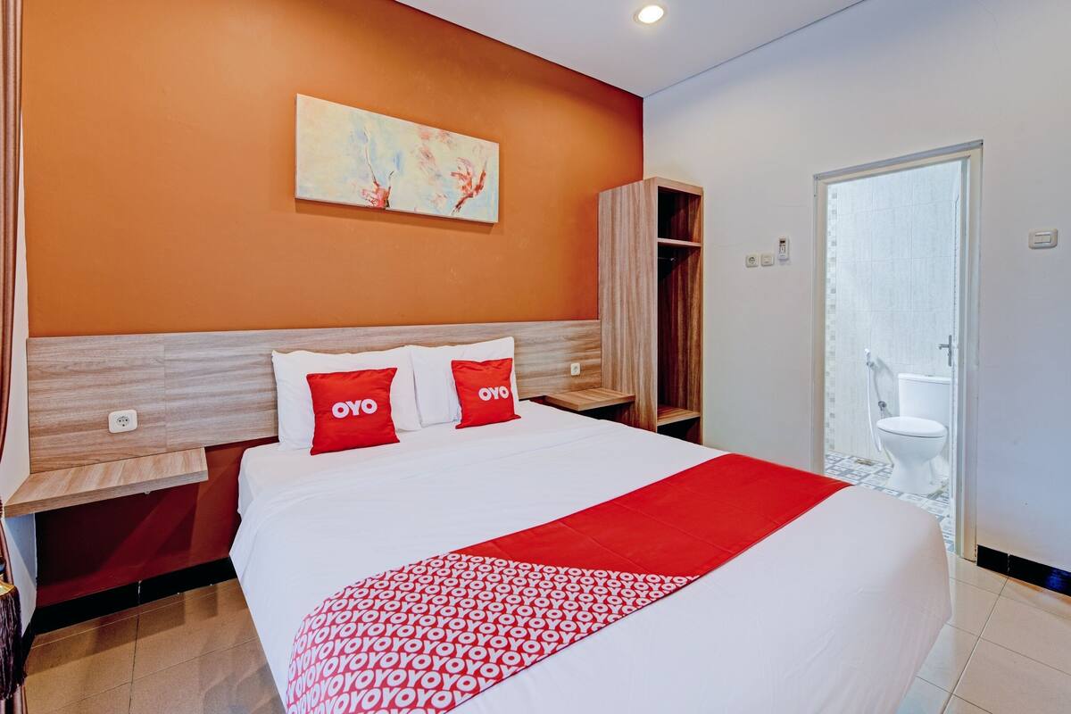 A hotel room with a large bed, a red and white bedspread, a wooden headboard, a wall-mounted painting, and a bathroom visible through an open door.