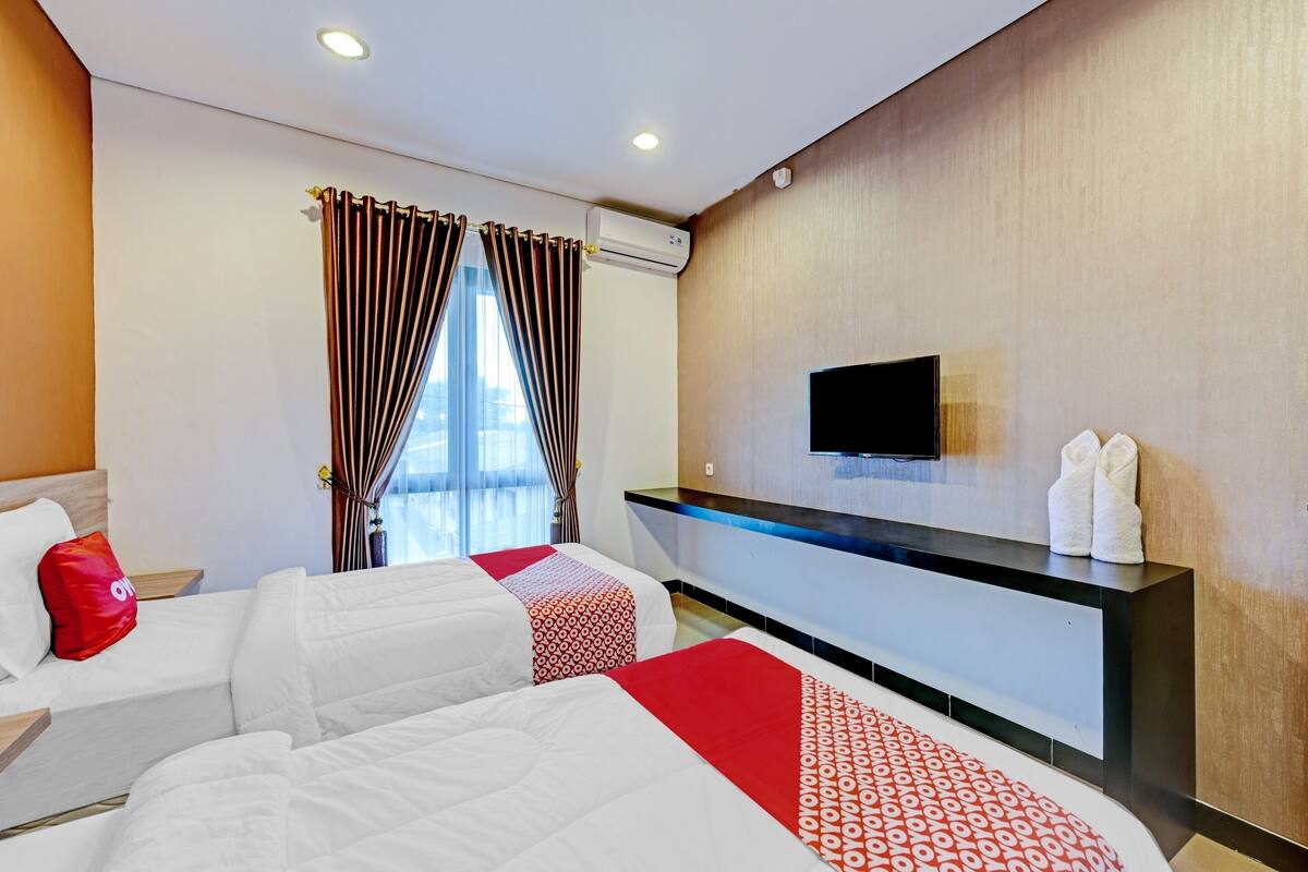 A hotel room with two beds, a flat-screen TV, a minibar, and a large window with curtains.