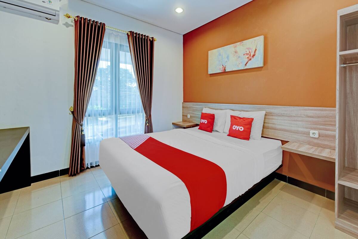 A hotel room with a bed, a nightstand, a painting on the wall, and a window with curtains.