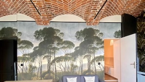 Una habitación con techo de ladrillo, un mural de árboles, un sofá gris con dos cojines y una mesa de madera.