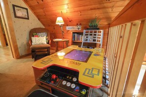 Une pièce avec un plafond en bois, une chaise avec un coussin, une petite table avec une lampe, une bibliothèque et une table de jeux.