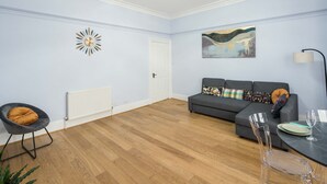 A living room with a grey sofa, a clear chair, a dining table with a plate and glass, a radiator, and a wall clock. - Gloucester Station Gem, 3 Bedroom Oasis (London)