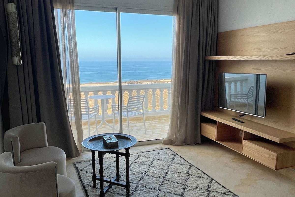 A hotel room with a bed, a TV, a seating area with a table, and a view of the sea.