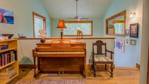 A room with a wooden piano, a chair, a bookshelf, a window, and a ceiling fan.