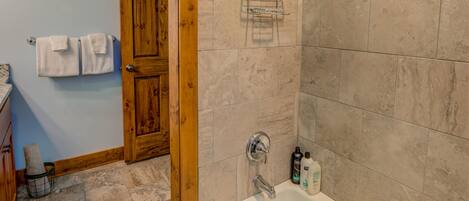 A bathroom with beige tiles, a wooden door, a shower with a glass shelf, and a sink with toiletries.