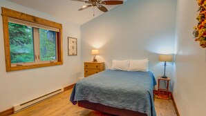 A bedroom with a bed, a ceiling fan, a window with a view of trees, and a nightstand with a lamp.