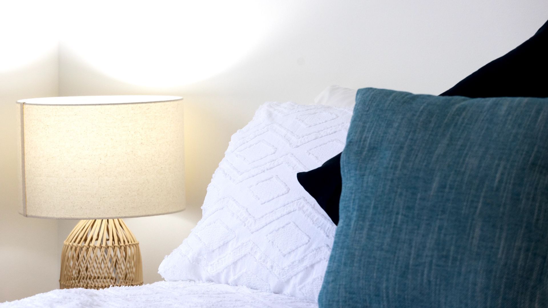 Ein ordentlich bezogenes Bett mit weißen und türkisen Kissen, einer weißen, strukturierten Tagesdecke und einem gewebten Nachttisch mit Lampe.