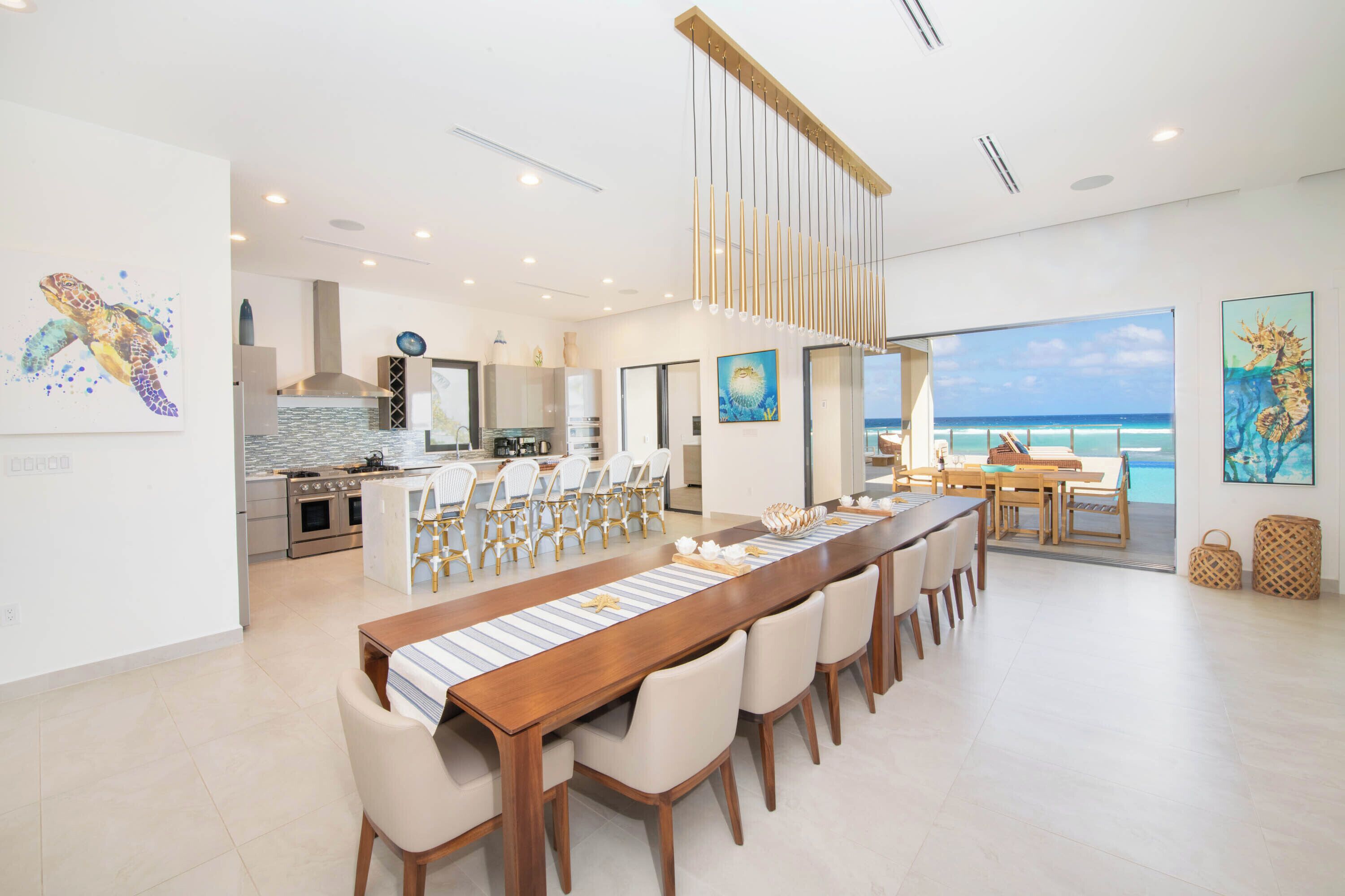 A spacious dining area with a long wooden table and multiple chairs, a kitchen in the background, and a view of the ocean through large windows.