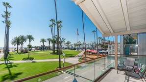 A balcony with a clear view of a beach, palm trees, and a flag. - 6BR Oceanfront Retreat, Steps to Beach & Balboa Pier with AC! (Newport Beach)